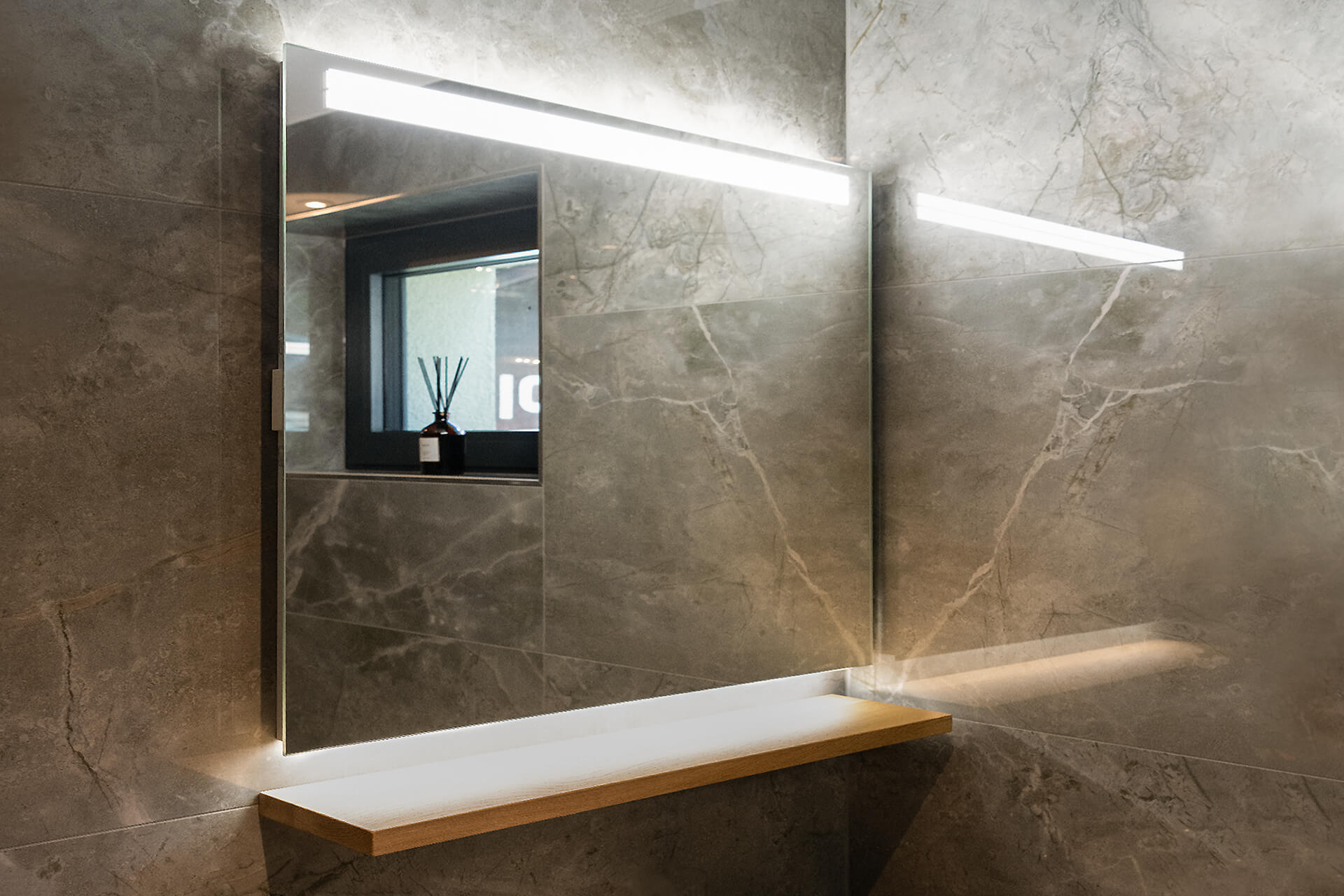 Miroir lumineux ARANGA Line LED - Des lampes LED de haute qualité, commutables individuellement, diffusent une lumière directe en haut et une lumière indirecte en bas. Miroir lumineux ARANGA Line LED monté sur un mur en plaques de céramique. Bandeau lumineux en haut et en bas. Tablette en bois.