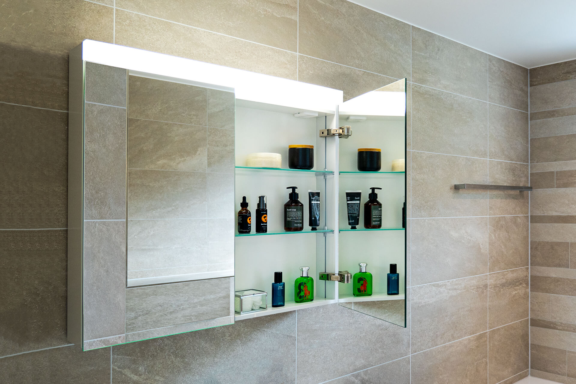 LOW Line BASIC LED Spiegelschrank - Oben leuchtet eine Leuchte den Raum optimal aus. LOW Line BASIC LED Spiegelschrank an Keramikplattenwand mit offener Türe. Duschtuchhalter.