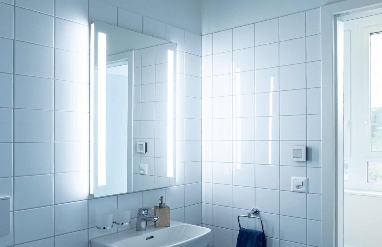 TRI Line LED Lichtspiegel - Ins Gästebad passt er ebenso gut wie in das Entrée. TRI Line LED Lichtspiegel an Keramikplattenwand. 2 seitliche Lichtbänder. Lavabo unten. Fenster auf rechter Seite.